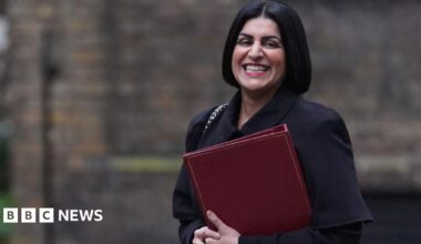 Shabana Mahmood is smiling as she walks in Downing Street. She is holding a red folder and wearing a dark coloured coat.