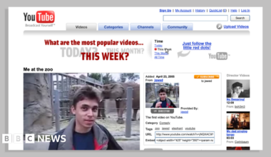 An early YouTube watchpage. The red and black YouTube logo is seen against a white web page background. The page features an image of Jawed Karim from his video 'Me at the Zoo'. Kawed is a young man with dark hair and is pictured standing in front of an elephant enclosure.