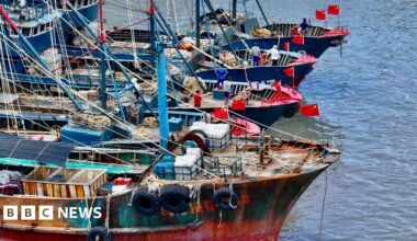 Four fishing vessels with arrays of lines and nets on their decks float in a line on calm water. They are all flying Chinese flags and three of the boats have crew standing near their bows.