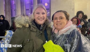 Robbie Williams singing into a microphone. He is wearing a jean shirt with a white collar and has grey spiky hair.