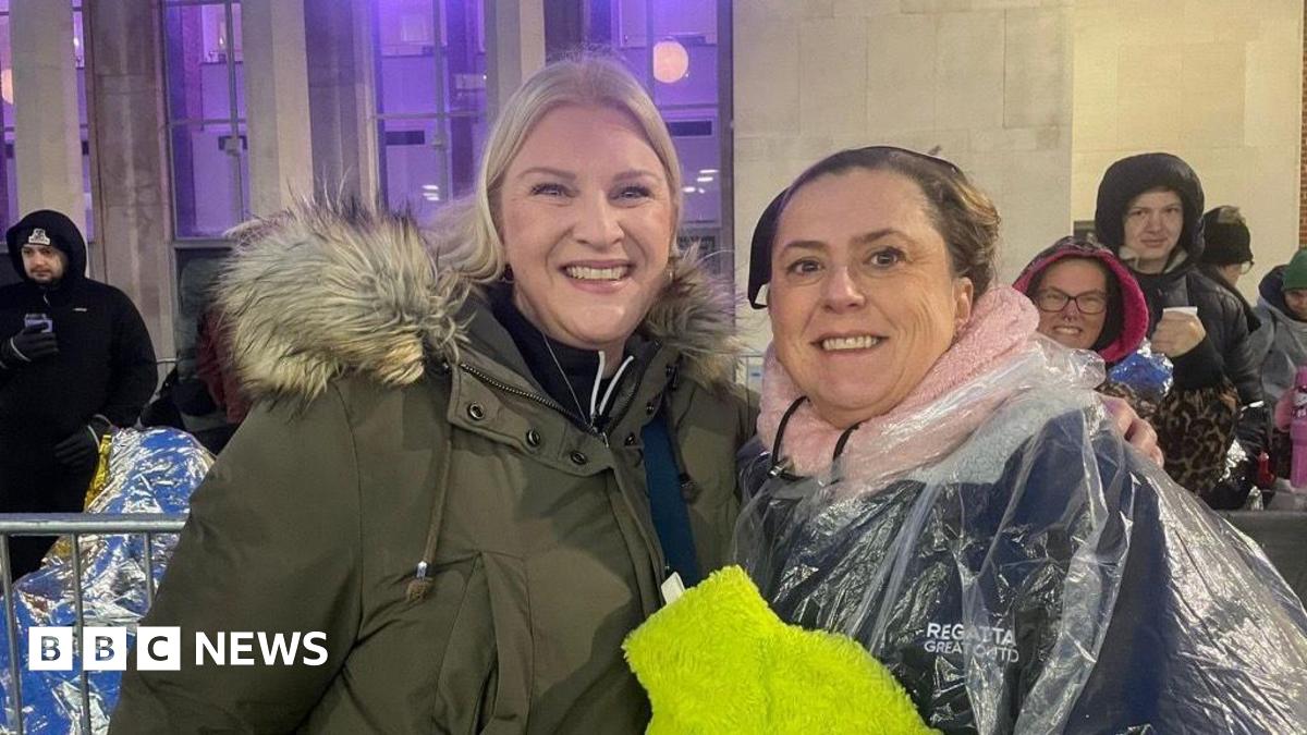 Robbie Williams singing into a microphone. He is wearing a jean shirt with a white collar and has grey spiky hair.
