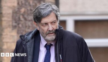 Alan Harris walking. He is wearing a pink top with a blue tie and blazer. He is wearing a black coat. The background shows a brown wall