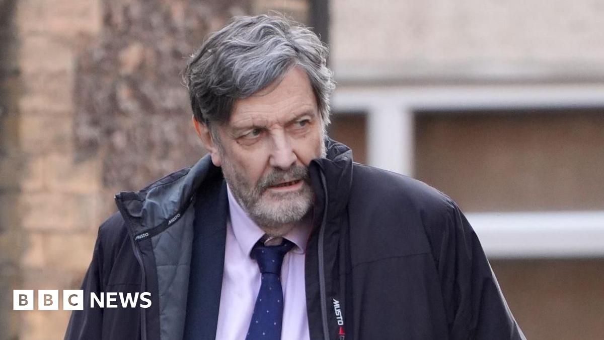 Alan Harris walking. He is wearing a pink top with a blue tie and blazer. He is wearing a black coat. The background shows a brown wall