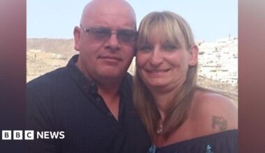 On the left, there is a man who is bald, wearing a black polo t-shirt with dark grey framed sunglasses. On the right, there is a woman in a dark blue off-the-shoulder t-shirt, with a tattoo next to her shoulder. She has blonde hair.