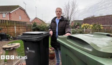 A lady in her seventies standing in a garden looking at the camera with two large wheelie bins next to her.