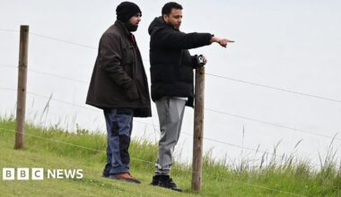 Walid Saadaoui (right) and Amar Hussein stand on a cliff edge, Saadaoui  leans against a wooden post in the ground that holds up a fence. He wears a black jacket, white t-shirt and grey jogging bottoms. Hussein wears a brown jacket and dark grey trousers