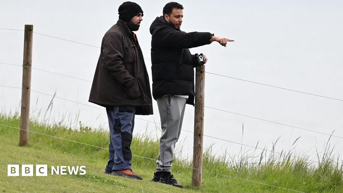 Walid Saadaoui (right) and Amar Hussein stand on a cliff edge, Saadaoui  leans against a wooden post in the ground that holds up a fence. He wears a black jacket, white t-shirt and grey jogging bottoms. Hussein wears a brown jacket and dark grey trousers