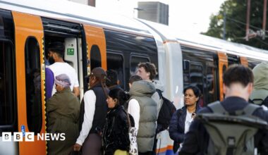 Passengers board a crowded London Overground train, with people queuing at the open doors of an orange-and-white carriage.