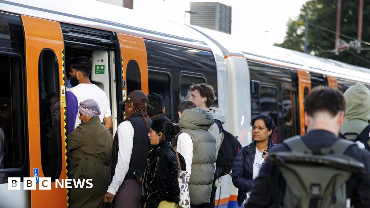 Passengers board a crowded London Overground train, with people queuing at the open doors of an orange-and-white carriage.