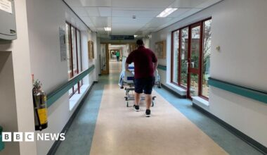 A man in a red t-shirt and shorts pushing a hospital bed through a hospital corridor, walking away. The walls are white, with blue hand rails through them, and large windows along the corridor.