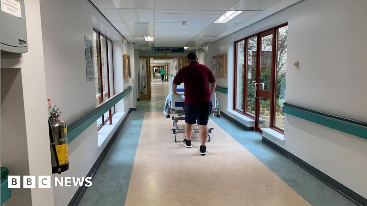 A man in a red t-shirt and shorts pushing a hospital bed through a hospital corridor, walking away. The walls are white, with blue hand rails through them, and large windows along the corridor.