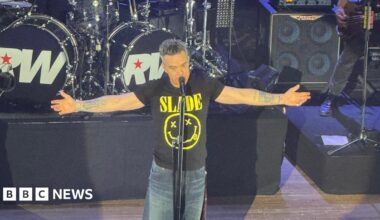 Robbie Williams in a black Slade tshirt is on stage with his arms outstretched at his sides
