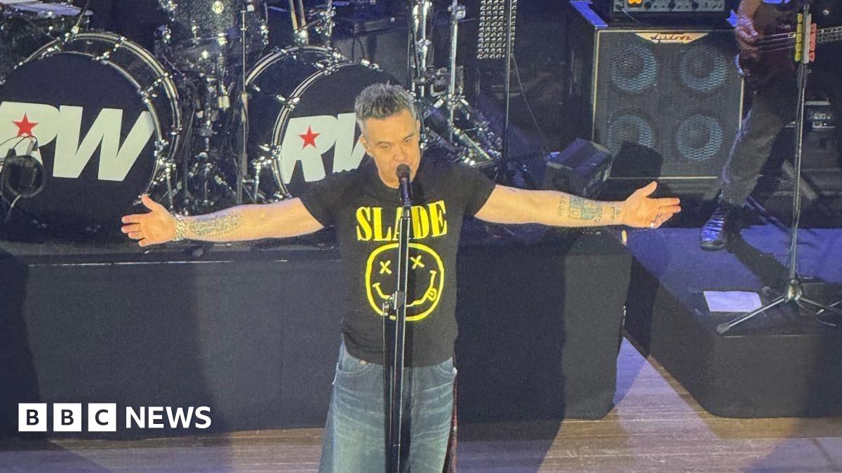 Robbie Williams in a black Slade tshirt is on stage with his arms outstretched at his sides