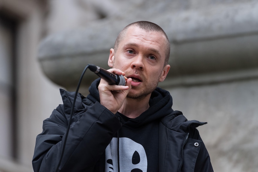 A man in his 30s is speaking while holding a mic.