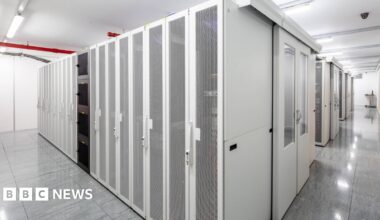 A corridor of white, mesh‑fronted server racks in a brightly lit data centre.