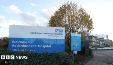 Exterior image of Addenbrooke's Hospital. In the foreground is a sign welcoming people to the hospital's Hills Road entrance. In the background are hospital buildings.