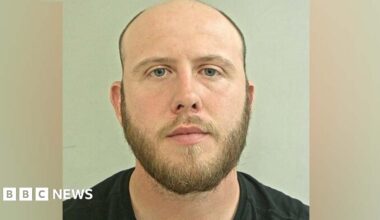 Peter Sayle's mugshot. He is bald on top of his head with very short dark hair at the sides and a short beard. He is wearing a black t-shirt against a plain grey background