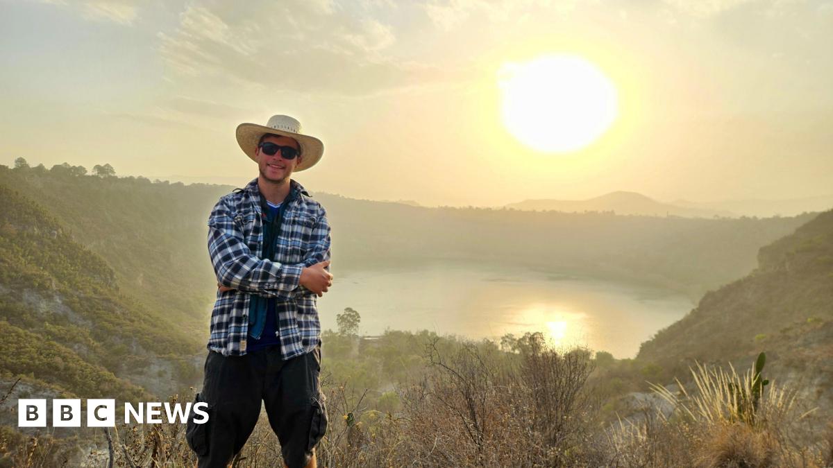Gus in Mexico, standing on a hill with his arms crossed in a chequered overshirt, sunglasses and a wide-brimmed sun hat with a lake and low sun in the background
