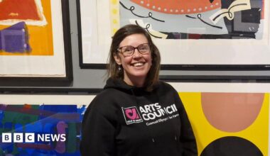 Suzanne, a woman with short brown hair, she wears glasses and has a bright smile as she stands in front of bright contemporary art.