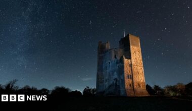 Orford Castle on a clear night. Stars can be seen in the sky above. The castle is lit by an orange glow.