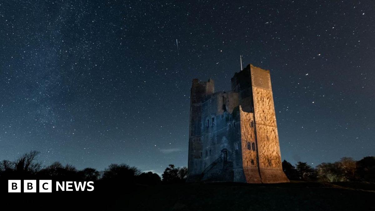 Orford Castle on a clear night. Stars can be seen in the sky above. The castle is lit by an orange glow.