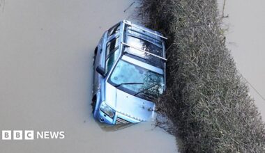 A silver Land Rover stuck in flood water. It is slightly tipped to the side and water is coming up over one of its headlights.