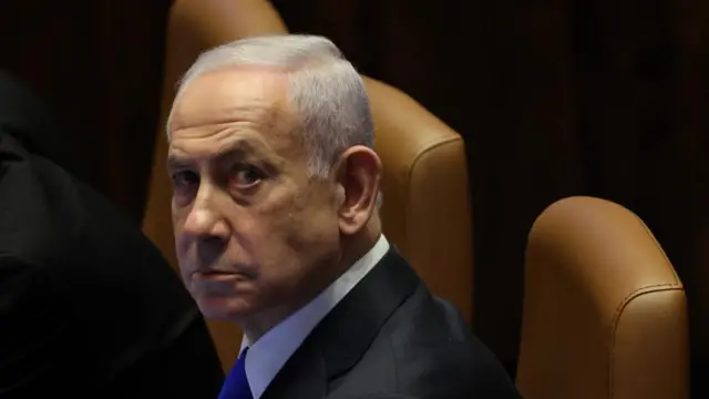 Benjamin Netanyahu, wearing a dark suit with a blue tie, looks up from his seat