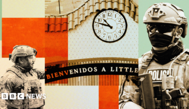 A man carries a box of groceries while crossing a street in a neighbourhood with many business signs in Spanish