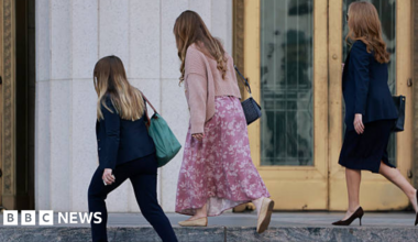 Three women seen from behind are approaching a building. One is shorter and wearing a dark trouser suit, the middle one is wearing a floaty dress and the one on the right is wearing a  dark skirt suit.