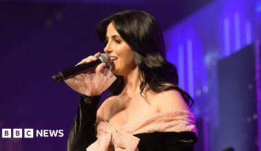 Lisa McHugh is singing into a black microphone. She is wearing a black dress with a nude poke a dot bow at the front. She has long black hair. The background is various shades of blue and purple.