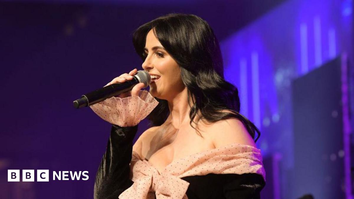 Lisa McHugh is singing into a black microphone. She is wearing a black dress with a nude poke a dot bow at the front. She has long black hair. The background is various shades of blue and purple.