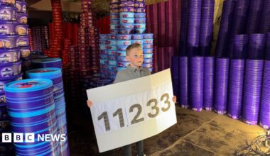 Teddy  holding a sign with the number 11,233 which reveals how many chocolate tubs he collected as part of his goal. He is surrounded by columns of colourful tubs and their lids that tower above him. He is smiling.