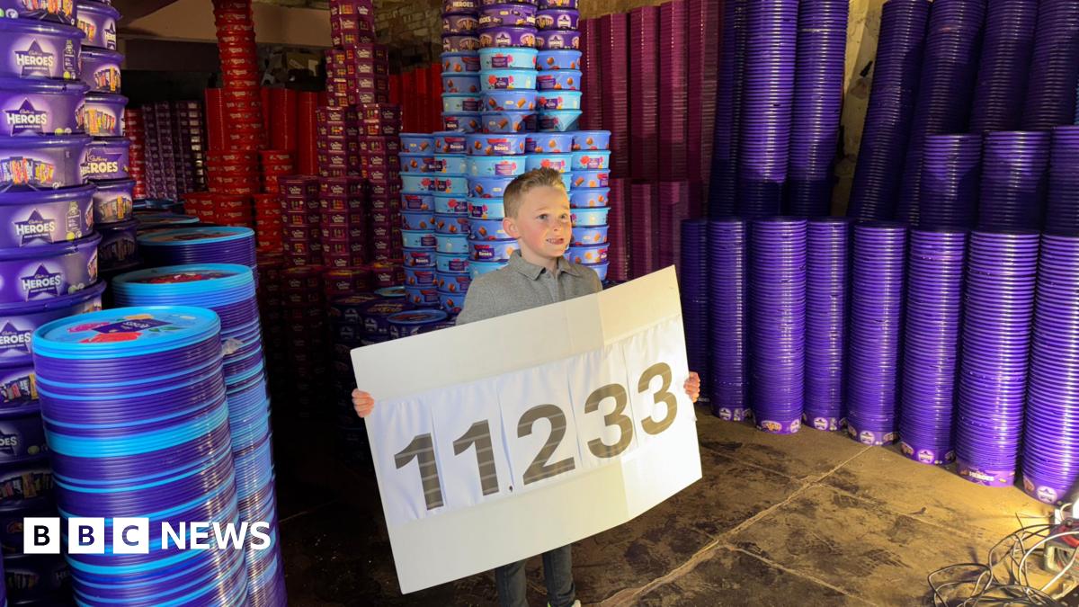 Teddy  holding a sign with the number 11,233 which reveals how many chocolate tubs he collected as part of his goal. He is surrounded by columns of colourful tubs and their lids that tower above him. He is smiling.