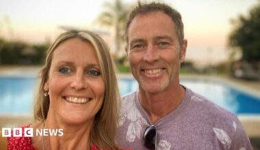 Lindsay, left, who has blonde mid length hair and is wearing a red dress, next to her husband Craig, who is wearing a mauve t-shirt with a white print. Both are smiling in the sun in front of a blue pool.
