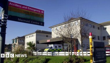 A general view of the Royal Cornwall Hospital in Truro on a sunny day. In the foreground is a sign pointing to Accident and Emergency, Link Corridors, Trelawny Wing and Mermaid Centre. There is an ambulance parked outside the main entrance to the building. To the right is a sign which reads: Warning when light flashes please cross over.