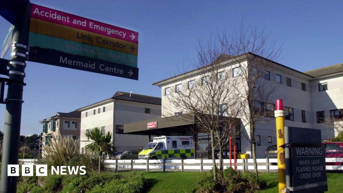 A general view of the Royal Cornwall Hospital in Truro on a sunny day. In the foreground is a sign pointing to Accident and Emergency, Link Corridors, Trelawny Wing and Mermaid Centre. There is an ambulance parked outside the main entrance to the building. To the right is a sign which reads: Warning when light flashes please cross over.