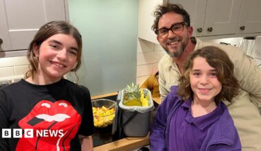 A father and two children stand smiling in their kitchen beside a bowl of fruit salad and their kitchen caddy filled with fruit peelings.