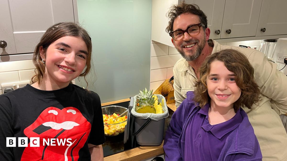 A father and two children stand smiling in their kitchen beside a bowl of fruit salad and their kitchen caddy filled with fruit peelings.
