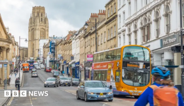 Park Street. It is a busy, hilled road with cars, busses and a cyclist travelling on it. The Wills Memorial building is at the top of it with a grand tower. Shops line the street and pedestrians walk up and down the pavement.