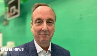 Ian Thorn smiles at the camera. He is wearing a blue jacket and check shirt. Behind him is a green wall