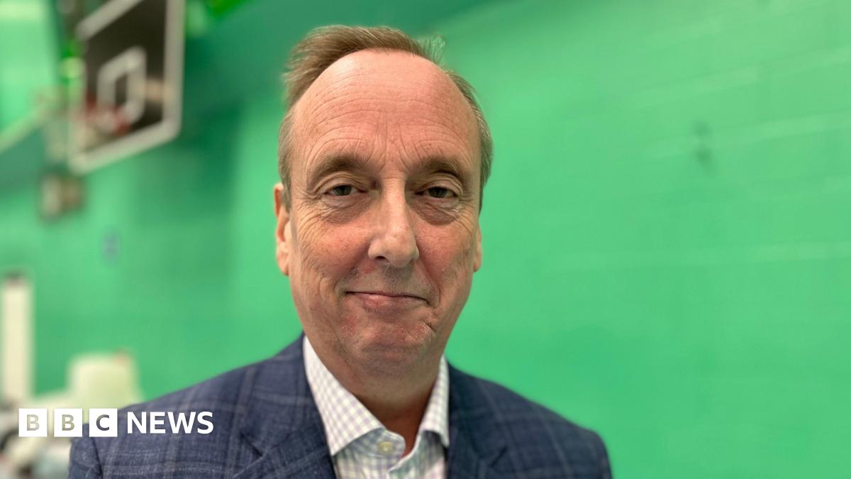 Ian Thorn smiles at the camera. He is wearing a blue jacket and check shirt. Behind him is a green wall