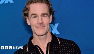 James Van Der Beek wears a black polo and stands in front of a blue background