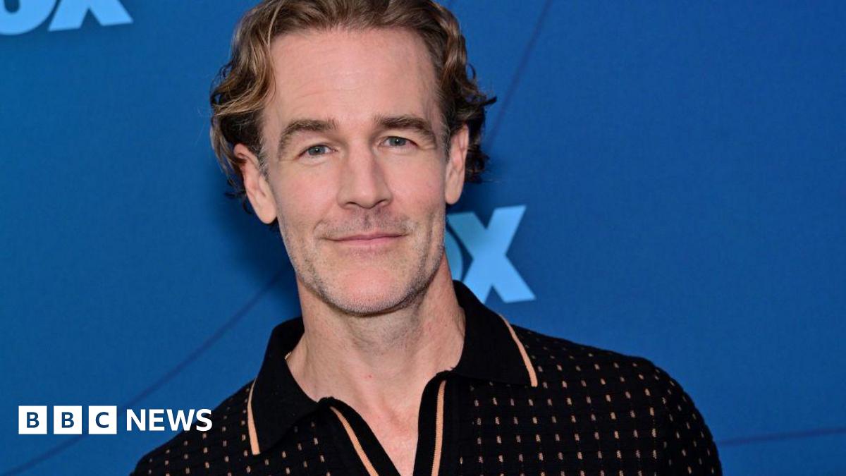 James Van Der Beek wears a black polo and stands in front of a blue background