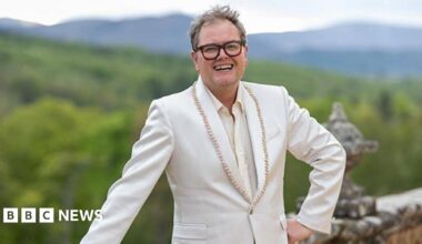 Alan Carr, a man with unkempt hair and thick-rimmed glasses. He is standing with one hand on his hip and the other leaning on a wall in front of lots of greenery. He is smiling which shows off a gap in his teeth. He is wearing a cream suit jacket with gold beading on it.