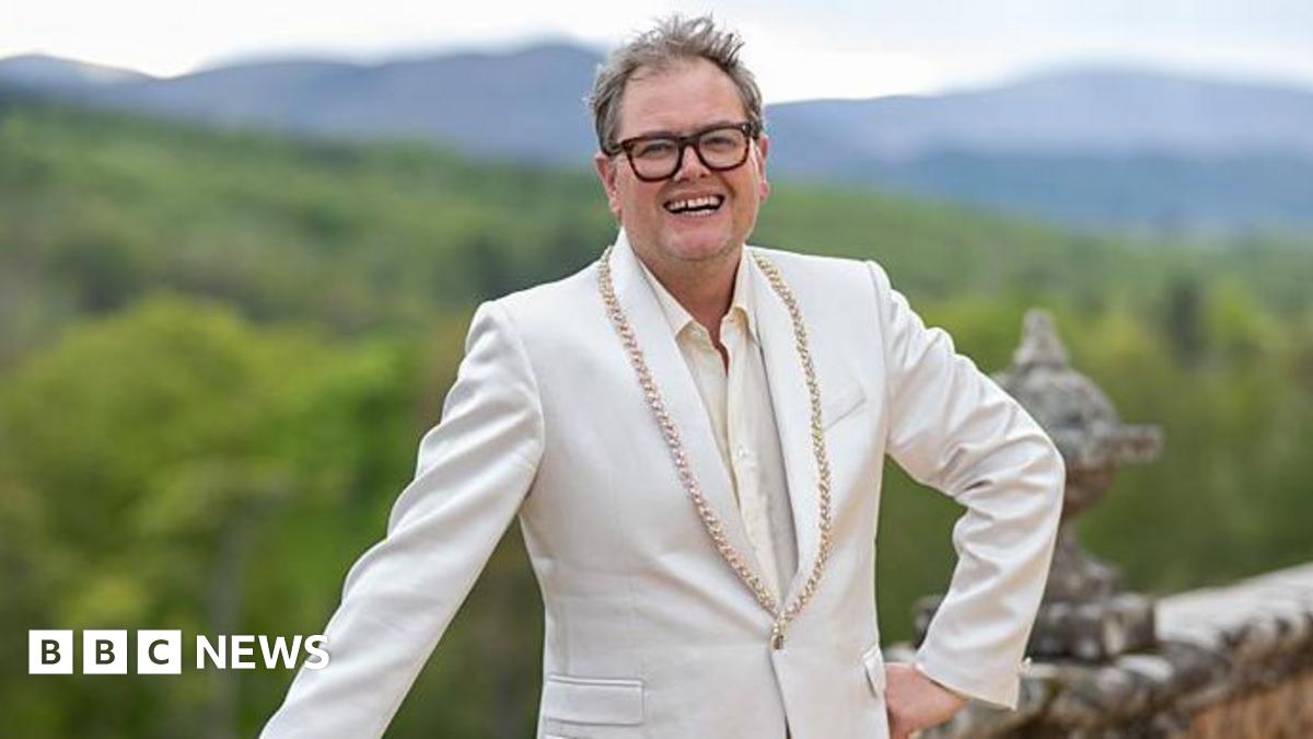 Alan Carr, a man with unkempt hair and thick-rimmed glasses. He is standing with one hand on his hip and the other leaning on a wall in front of lots of greenery. He is smiling which shows off a gap in his teeth. He is wearing a cream suit jacket with gold beading on it.