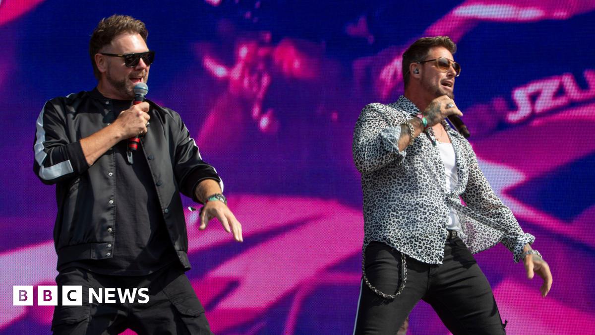 Two male singers stand on a large concert stage in front of a bright pink and purple LED screen. Both wear sunglasses and hold handheld microphones. One is dressed in a black jacket and black trousers, while the other wears a patterned shirt over a white top with black trousers and a chain at his waist. Stage lighting and part of a band setup are visible in the background.