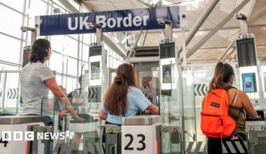 People approach electronic passport scanners at the UK border at Stansted airport in 2018 - one person wears an orange backpack and all have their backs to the camera. In the background is a sign saying 'UK Border'.