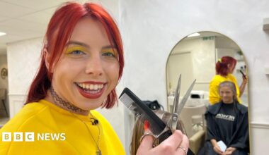 A woman smiling at the camera. She has bright red, shoulder-length hair, half tied back. She wears bright yellow eye shadow, which matches her bright yellow jumper. She is holding a black comb in her hand and silver scissors. Behind her a woman can be seen in a mirror wearing a black barber's apron an sitting in a chair having her hair cut.