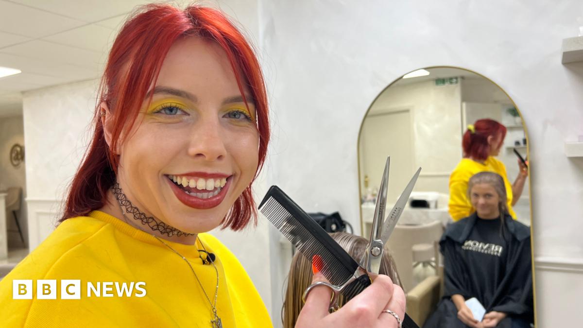A woman smiling at the camera. She has bright red, shoulder-length hair, half tied back. She wears bright yellow eye shadow, which matches her bright yellow jumper. She is holding a black comb in her hand and silver scissors. Behind her a woman can be seen in a mirror wearing a black barber's apron an sitting in a chair having her hair cut.