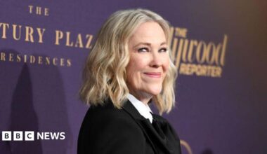 A woman with shoulder-length blonde hair is pictured in front of a purple backdrop. She wears a black suit jacket and a white shirt.